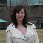 Woman with brown hair wearing a white jacket stands outdoors on a grassy area, with trees and buildings in the background.