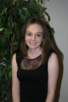 Young woman with straight brown hair wearing a sleeveless black dress stands indoors in front of a green leafy plant, smiling at the camera.