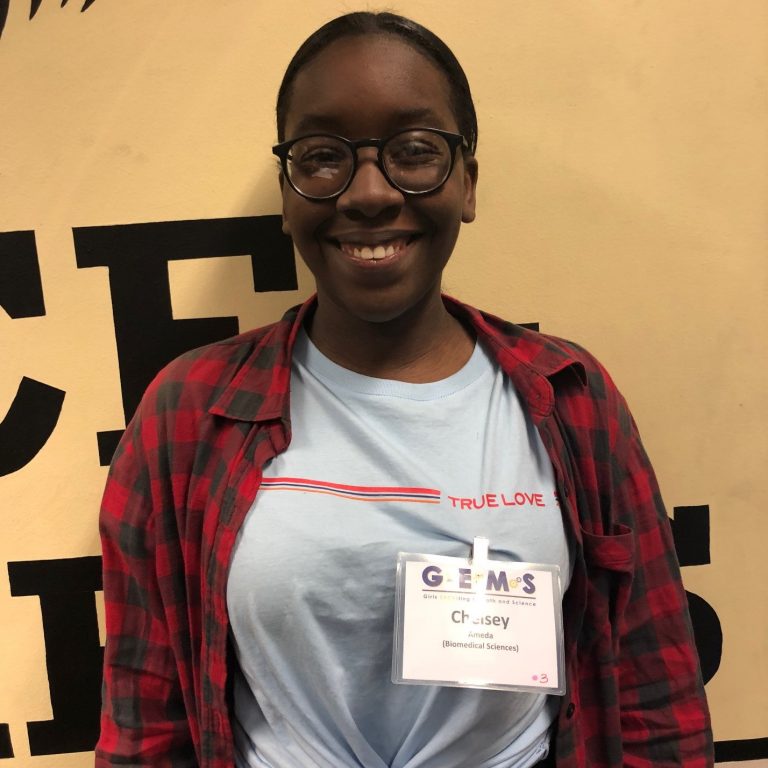 A person wearing glasses, a red plaid shirt, and a name badge labeled "GEMS" stands in front of a beige wall with black lettering.