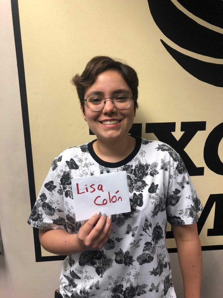 Person wearing glasses and a white floral shirt holds a paper sign that reads "Lisa Colón" while standing in front of a beige and black wall.