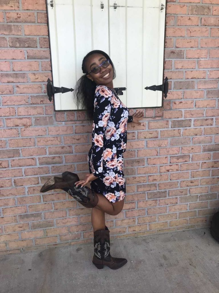 Woman in glasses and a floral dress poses playfully with one leg up, wearing cowboy boots, in front of a brick wall with a white shutter.