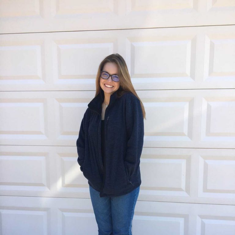 A person with long hair and glasses stands in front of a white garage door, wearing a dark jacket and blue jeans, smiling with hands in pockets.