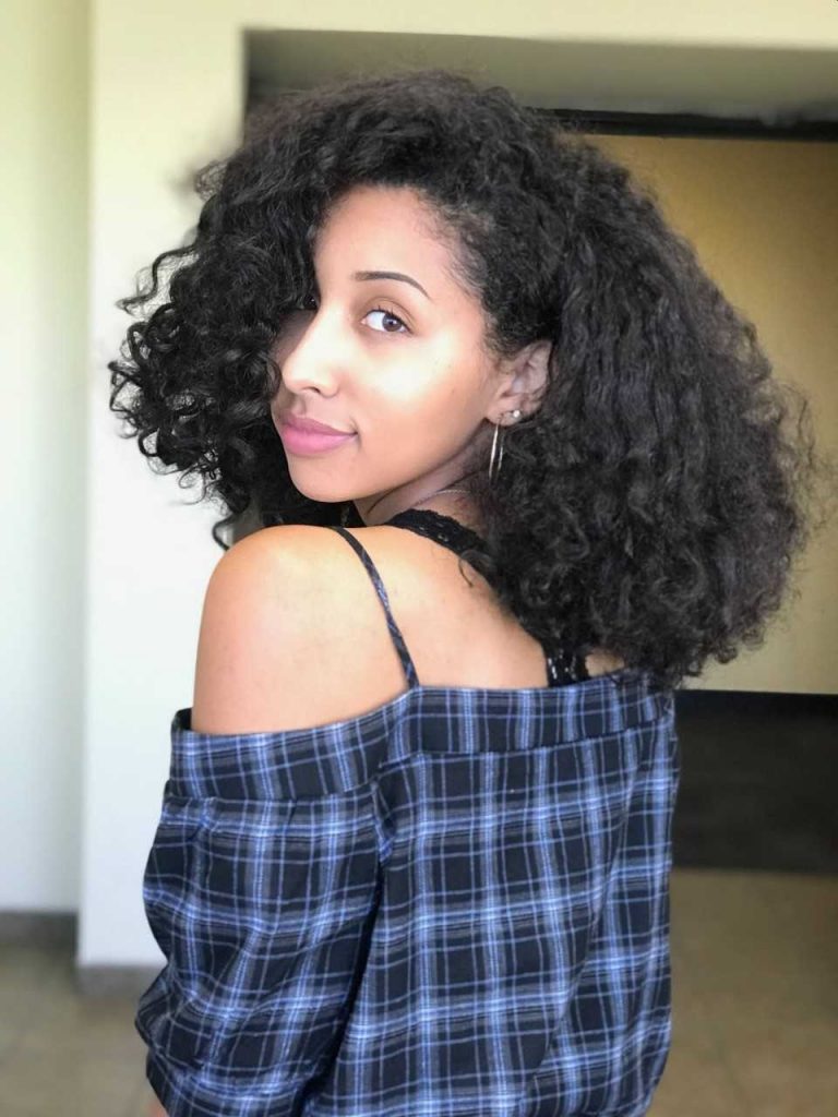 Woman with curly hair wears a blue plaid off-shoulder top, standing indoors and looking over her shoulder at the camera.