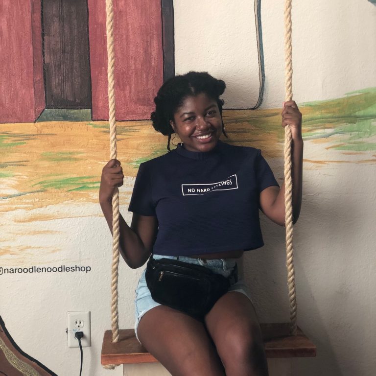 A woman with natural hair in twists sits on an indoor wooden swing, smiling, wearing a navy crop top and denim shorts. A painted mural is on the wall behind her.