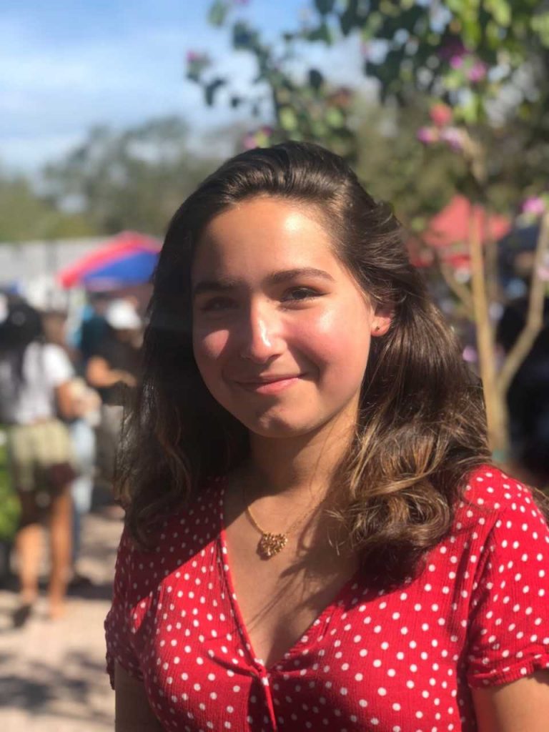 A young woman with wavy brown hair and a red polka dot shirt stands outdoors, smiling at the camera. Blurred people and trees are visible in the background.