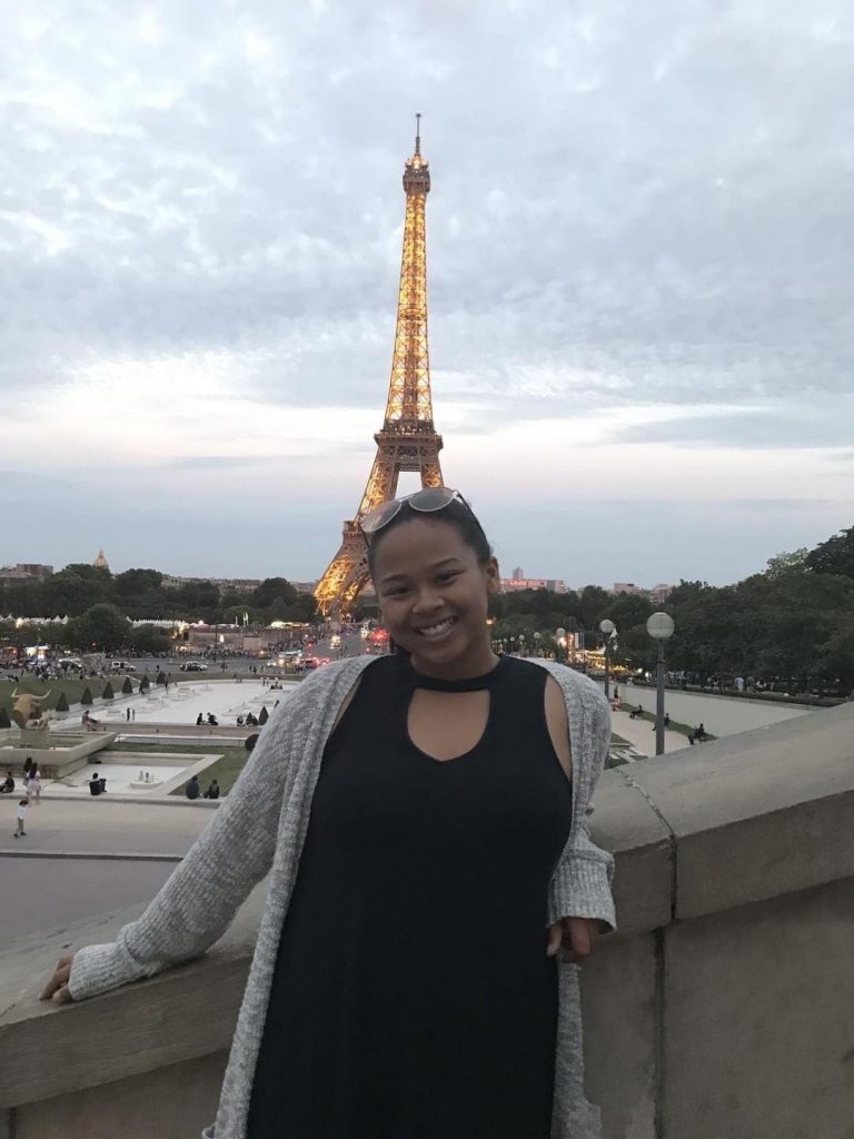 A person in a black dress and gray cardigan smiles in front of the illuminated Eiffel Tower at dusk in Paris.