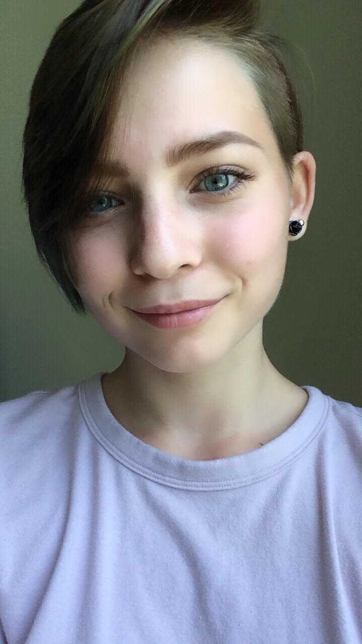 Person with short, side-swept brown hair, light skin, blue eyes, and wearing a pale pink shirt and black stud earrings, smiling at the camera against a neutral background.