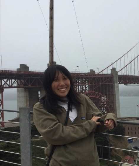 A smiling person in a jacket stands in front of the Golden Gate Bridge on a cloudy day.