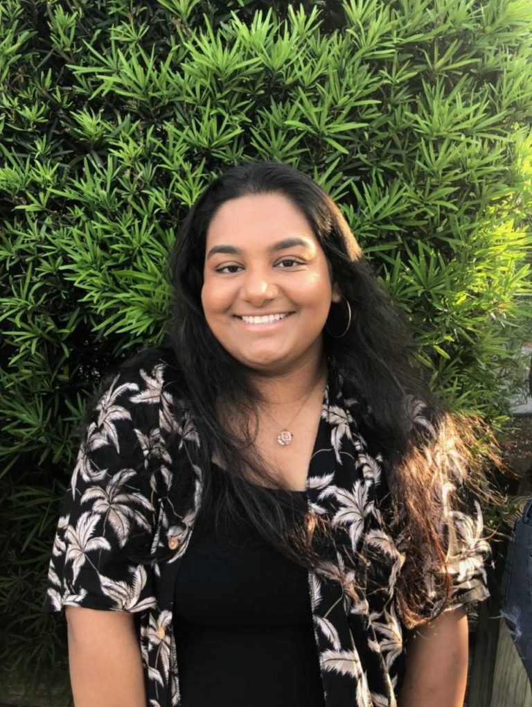 A woman with long dark hair, wearing a black top and patterned shirt, stands and smiles in front of green leafy bushes.