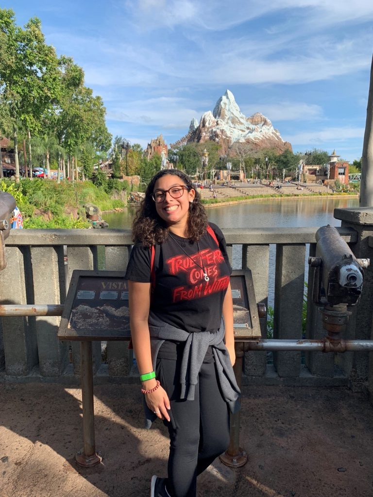 A person with long curly hair and glasses stands smiling on a bridge at a theme park, with a mountain attraction and trees visible in the background.