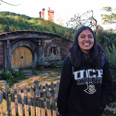 A woman wearing a UCF Knights hoodie stands smiling in front of a round wooden door and a grassy-roofed house resembling a Hobbit home.