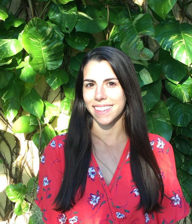 Woman with long dark hair, wearing a red floral blouse, stands in front of green leafy plants, smiling at the camera in bright sunlight.