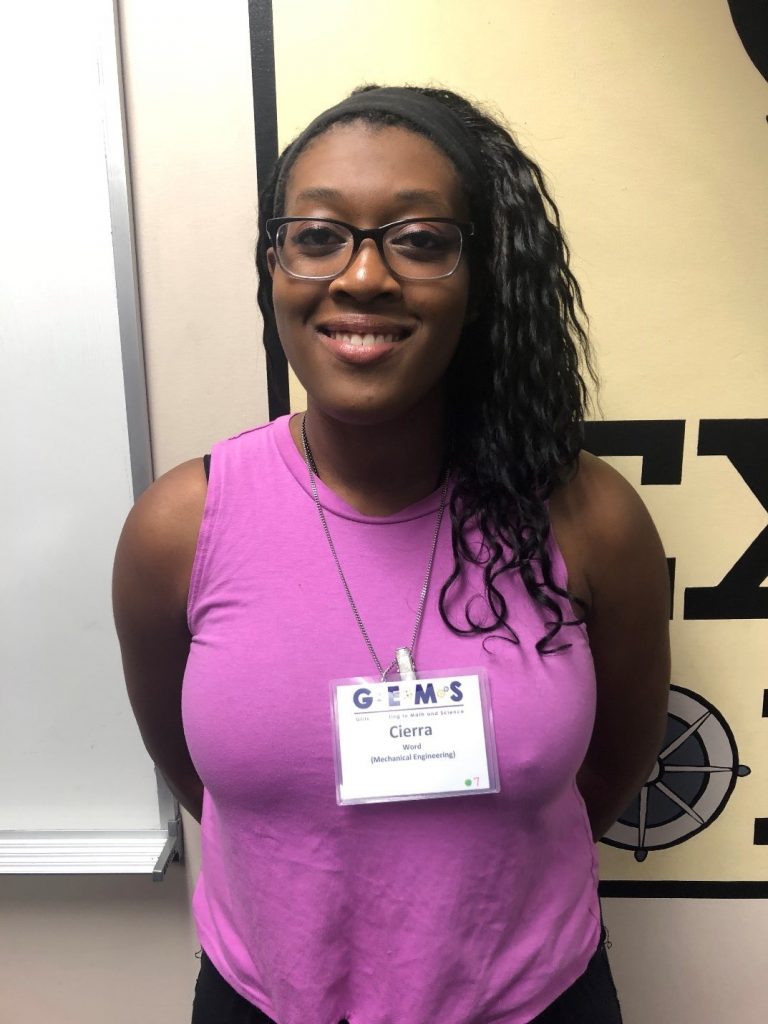 A woman wearing glasses and a purple sleeveless top stands indoors, smiling at the camera. She has a conference name badge that reads "Cierra" and "Mechanical Engineering.