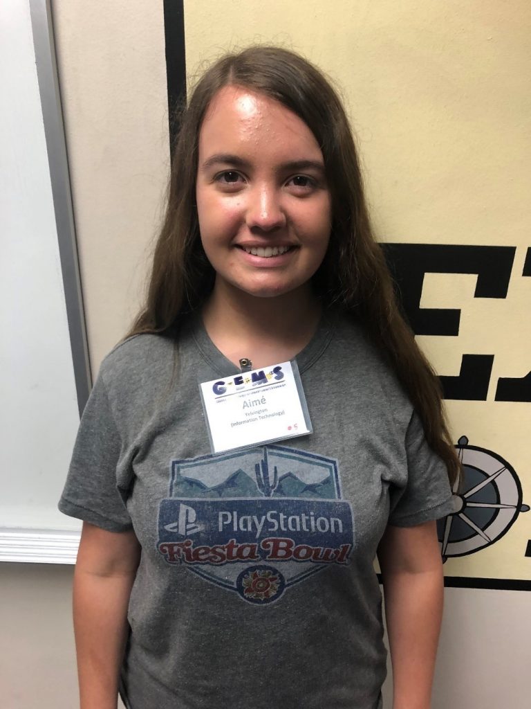 A young woman with long brown hair stands indoors, wearing a grey PlayStation Fiesta Bowl t-shirt and a name badge labeled "Alina.