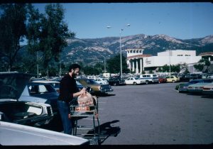 A man loads groceries into the trunk of a car in a parking lot with mountains and a Vons supermarket in the background.