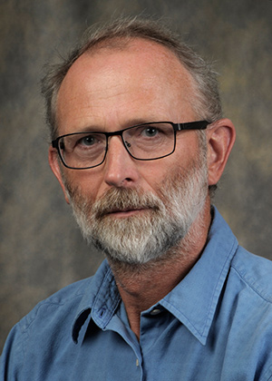 A middle-aged man with glasses, short gray hair, and a beard, wearing a blue collared shirt, posing against a neutral studio background.