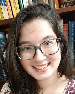 A young woman with long brown hair and glasses smiles at the camera, with bookshelves filled with books in the background.