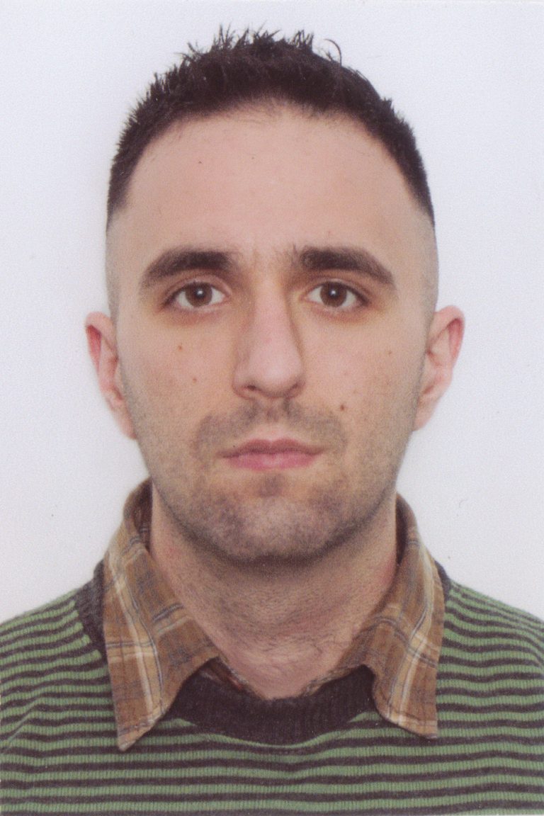 A man with short dark hair wearing a green striped sweater over a brown plaid shirt, looking straight at the camera with a neutral expression.