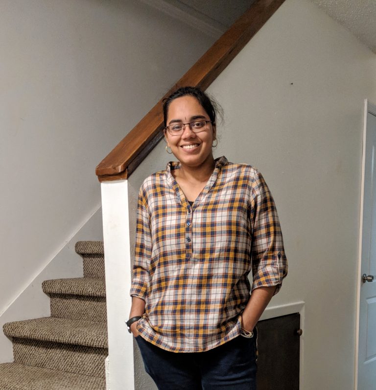 A person wearing glasses and a plaid shirt stands smiling in front of a staircase indoors.
