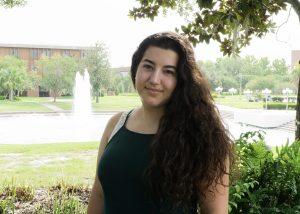 A woman with long curly hair stands outdoors in front of a pond with a fountain, trees, and a brick building in the background.