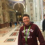 A woman with long wavy brown hair, wearing a black blazer and light-colored top, poses in front of a plain blue background.