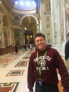 A person wearing a "Universita Roma" sweatshirt and tour badge smiles inside a large, ornate hall with marble floors and decorated walls and ceilings.