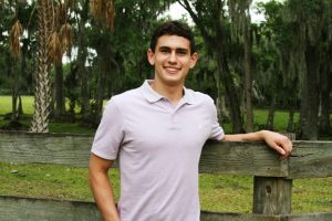 Young man in a light purple polo shirt stands outdoors, leaning on a wooden fence with trees and greenery in the background.