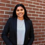A woman in a black blazer and polka dot blouse stands smiling in front of a brick wall.