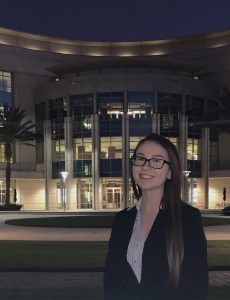 A woman in business attire stands smiling in front of a large, modern building at night, with lights illuminating the entrance and palm trees on either side.