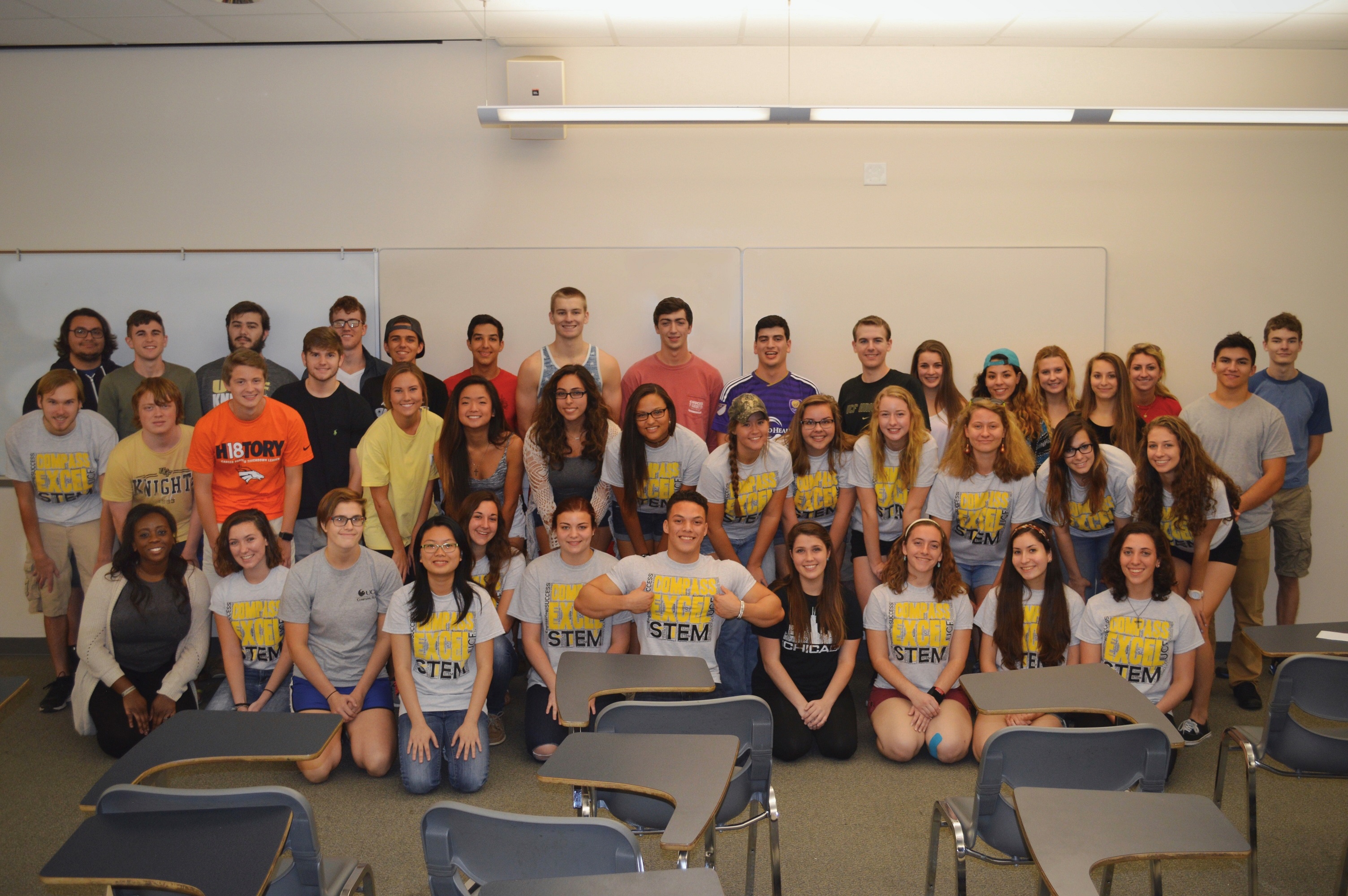 A group of students poses in a classroom, some seated and some standing, with several wearing matching STEM-themed t-shirts.