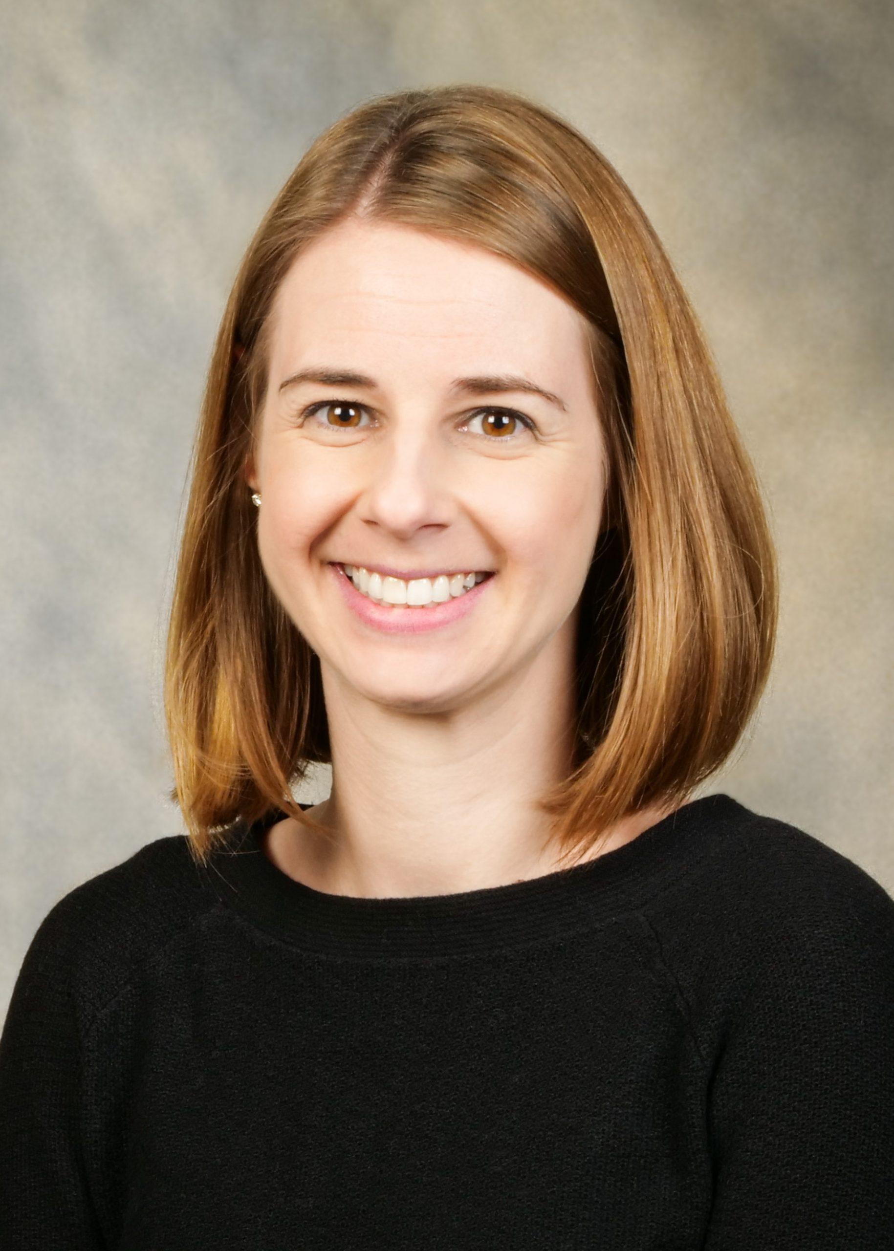 A woman with straight, shoulder-length light brown hair smiles at the camera. She is wearing a black top and is posed against a neutral, softly blurred background.