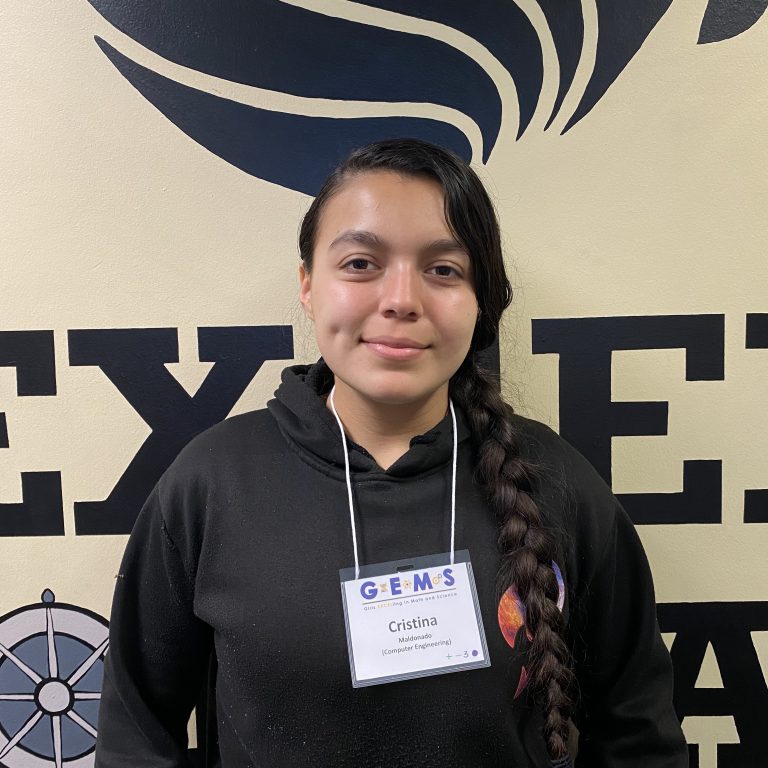 A young woman wearing a black hoodie and a GEMS name tag stands in front of a beige wall with bold text and graphic designs.