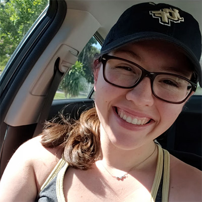 A woman wearing glasses and a dark cap smiles while sitting in a car with sunlight coming through the window.