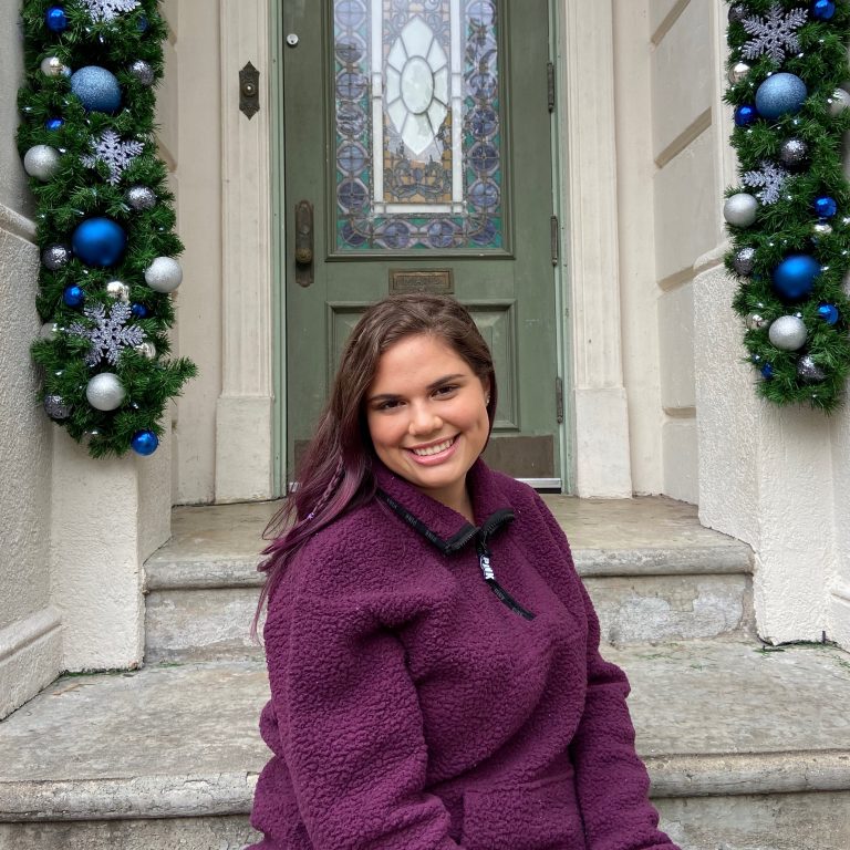 A woman in a purple sweater sits on stone steps in front of a green door decorated with two holiday garlands featuring blue, silver, and white ornaments.