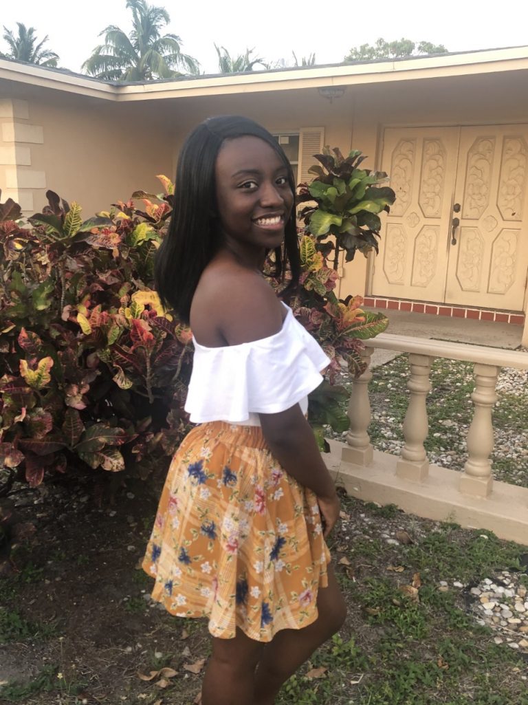 A woman stands outdoors by colorful shrubs in front of a house, wearing a white off-the-shoulder top and a yellow floral skirt, smiling at the camera.