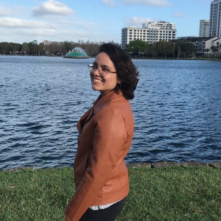 A person wearing glasses and a brown jacket stands by a lake with buildings and a fountain visible in the background.