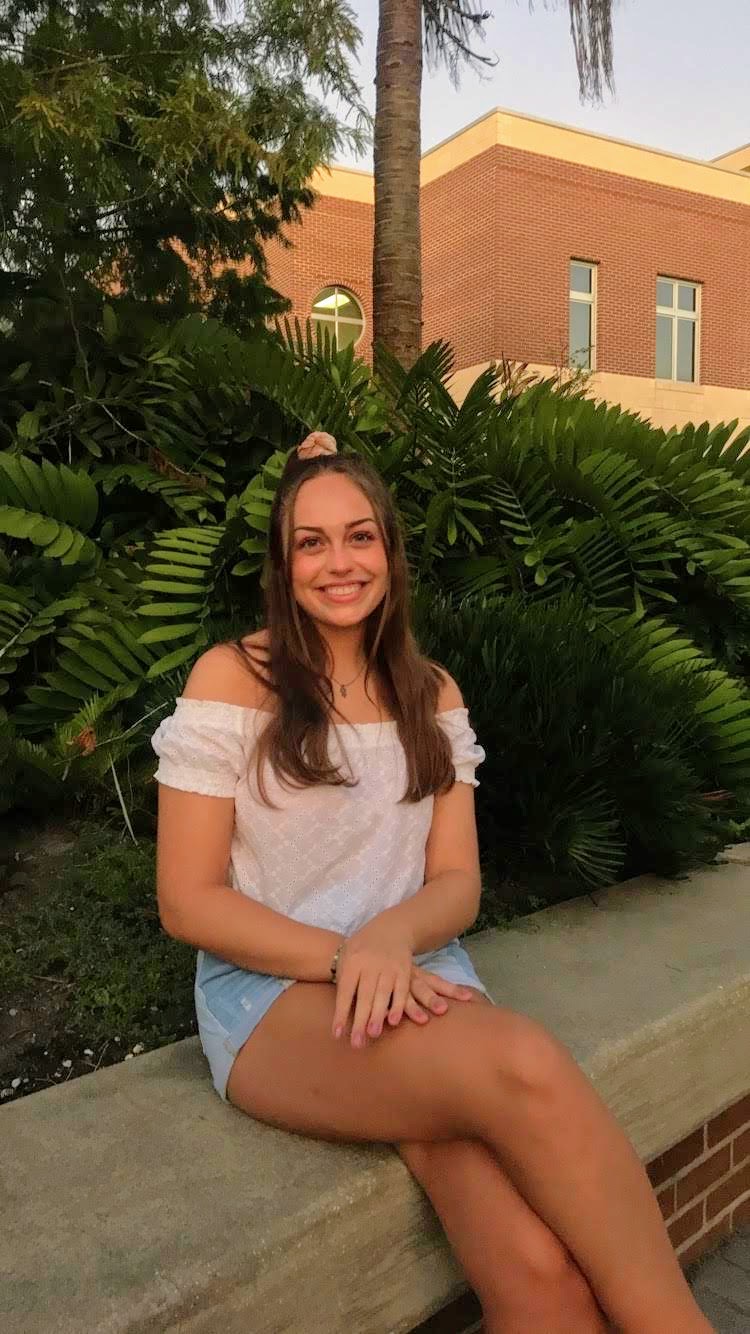 A young woman sits on a low concrete wall in front of green plants and a brick building, smiling at the camera.