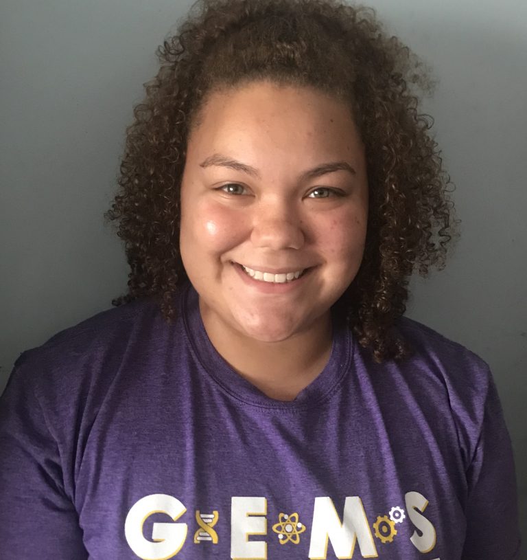 A person with curly hair smiles at the camera, wearing a purple shirt that reads "GEMS" with science-themed graphics.