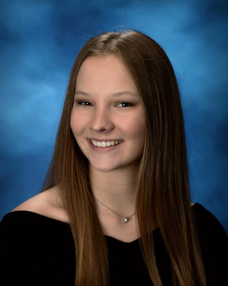 A young woman with long straight brown hair and a black off-the-shoulder top poses smiling in front of a blue studio background.