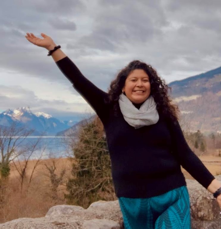 A person stands outdoors with one arm raised, smiling in front of a scenic landscape with mountains, a lake, and cloudy skies.