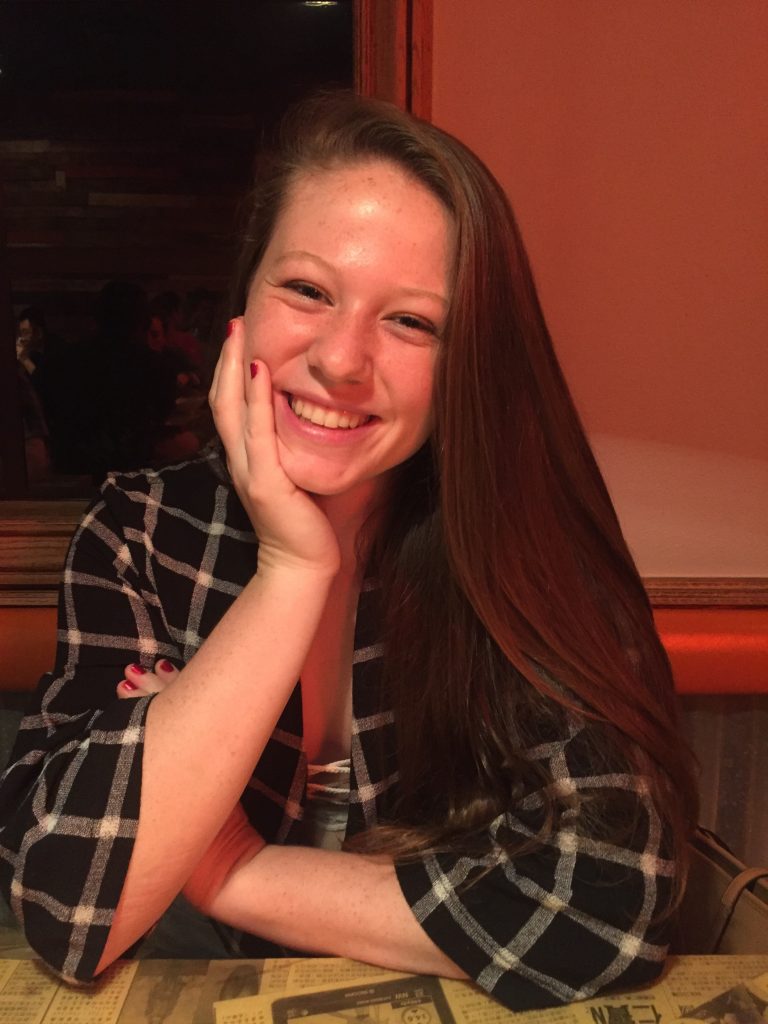 A young woman with long brown hair sits at a table, smiling with her head resting on her hand, wearing a black and white plaid shirt.