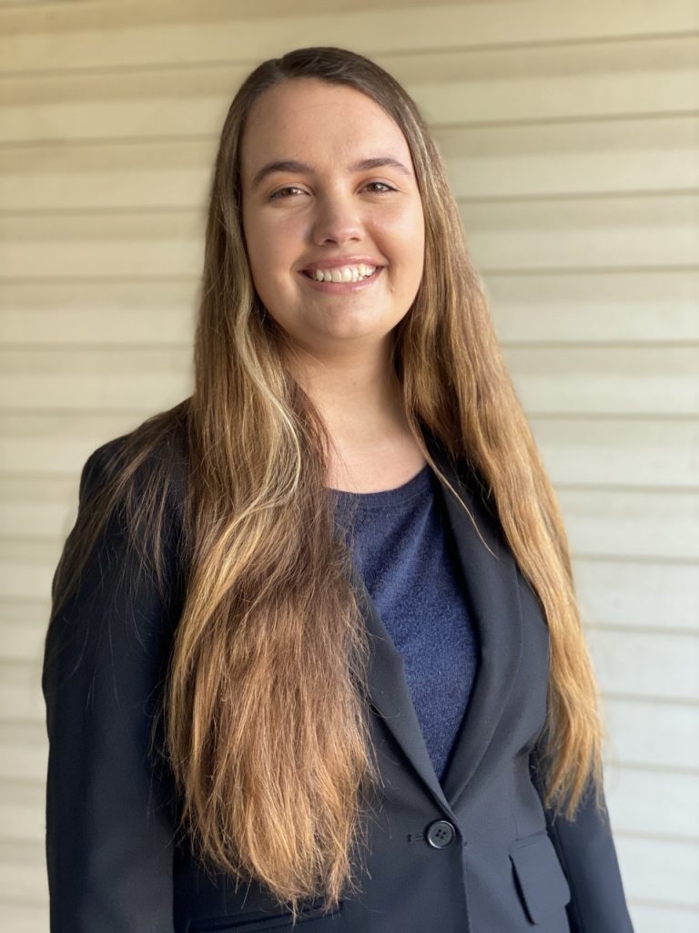 A young woman with long brown hair, wearing a dark blazer and navy top, stands in front of a light-colored wooden wall, smiling at the camera.