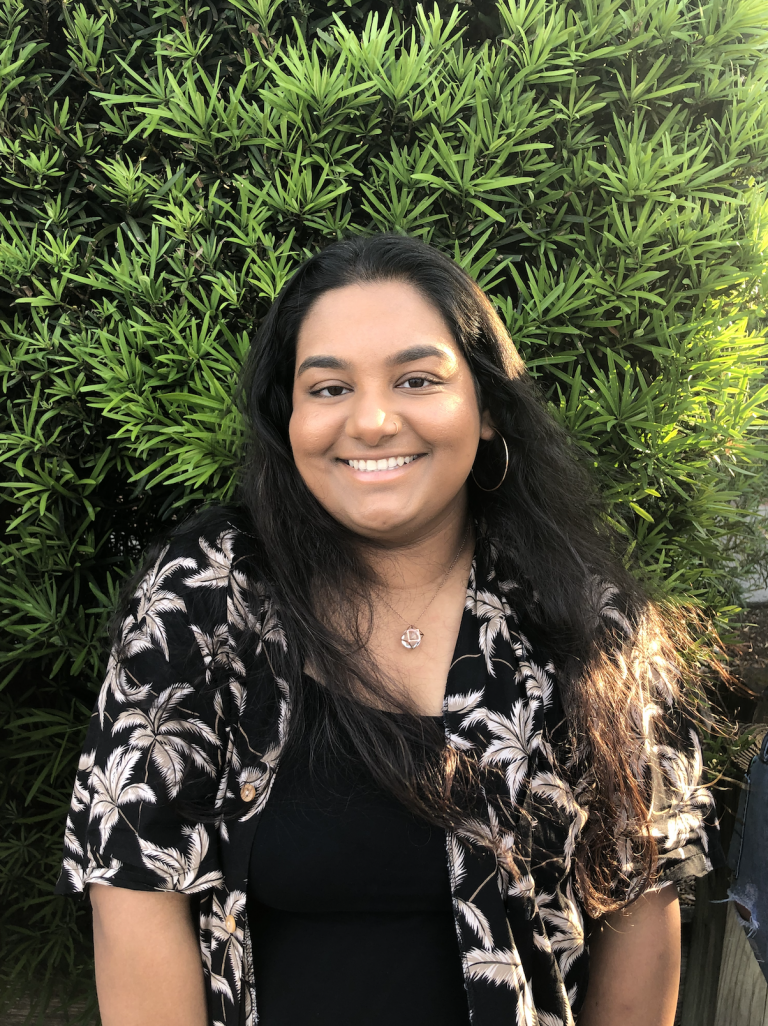 A person with long dark hair, wearing a black shirt and patterned shirt over it, smiles while standing in front of dense green foliage.