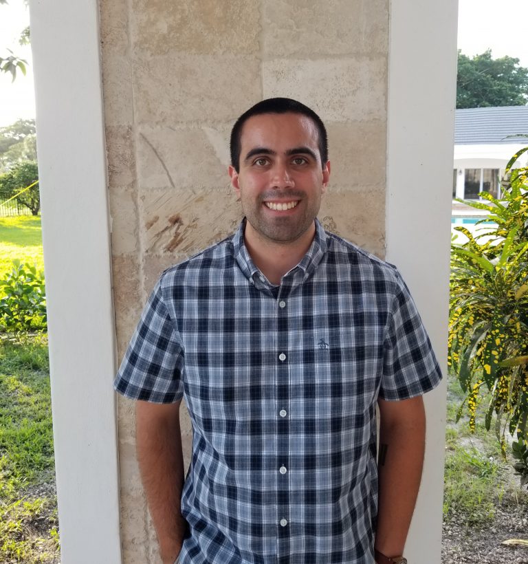A man wearing a blue and gray plaid short-sleeve shirt stands smiling in front of a stone wall outdoors.