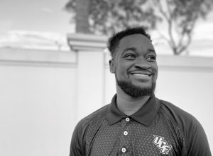 A man wearing a UCF polo shirt stands outdoors, smiling, with a wall and trees in the background. The image is in black and white.