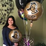 A woman stands indoors holding a UCF balloon, with more balloons nearby, next to a table and a decorative wall piece in the background.