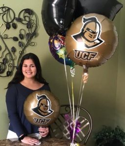 A woman stands indoors holding a UCF balloon, with more balloons nearby, next to a table and a decorative wall piece in the background.