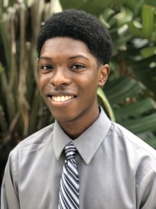 A young man wearing a gray dress shirt and striped tie smiles at the camera, standing outdoors in front of green leafy plants.