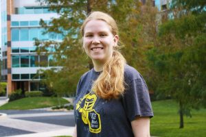 A young woman with blond hair in a ponytail stands outside on a grassy area, smiling, wearing a dark UCF Broad Cancer Center t-shirt, with trees and a modern building in the background.