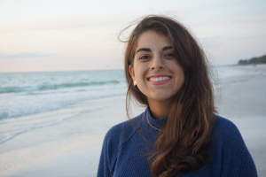A woman with long brown hair, wearing a blue sweater, smiles while standing on a beach at sunset.